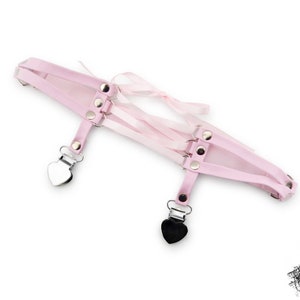 Adjustable pink thigh garter featuring multiple straps, silver heart-shaped suspender clips, and silver metal rivets. This cute thigh garter features a corset lacing design in the centre with adjustable ribbon ties for a secure fit, blending style with functionality. Perfect for alternative fashion enthusiasts, cosplay outfits, or making bold fashion statements. An edgy addition to pastel goth or kawaii style, combining a touch of romance with daring aesthetics. Displayed on a white background.