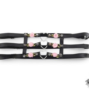 Black thigh garter featuring 3 horizontal straps with central silver heart ring hardware, pink satin roses and silver spike studs either side. Adjustable elastic back ensures a comfortable and versatile fit. Perfect for alternative fashion styles including pastel goth, Harajuku fashion and coquette aesthetics, as well as cosplay and BDSM communities. Displayed on a white background.