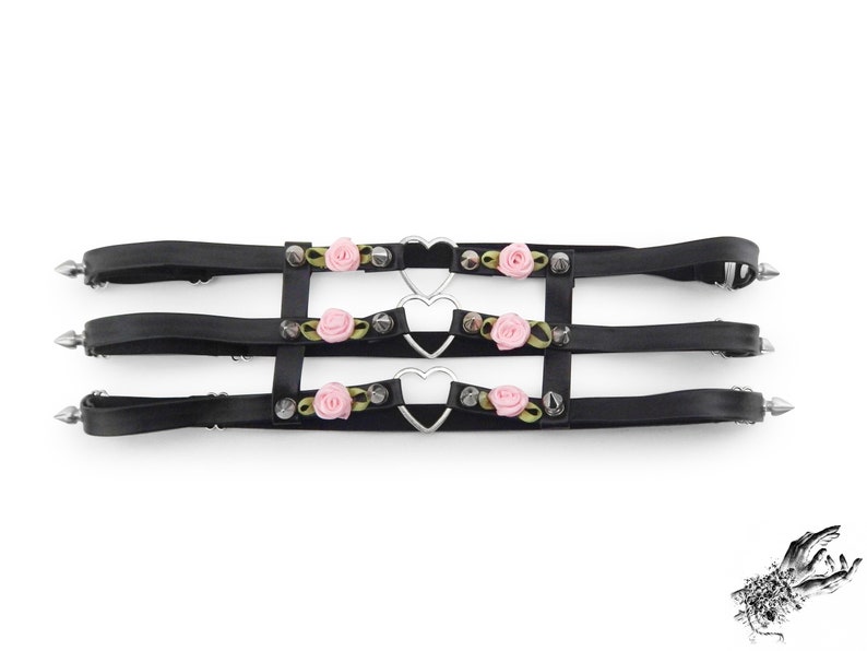 Black thigh garter featuring 3 horizontal straps with central silver heart ring hardware, pink satin roses and silver spike studs either side. Adjustable elastic back ensures a comfortable and versatile fit. Perfect for alternative fashion styles including pastel goth, Harajuku fashion and coquette aesthetics, as well as cosplay and BDSM communities. Displayed on a white background.