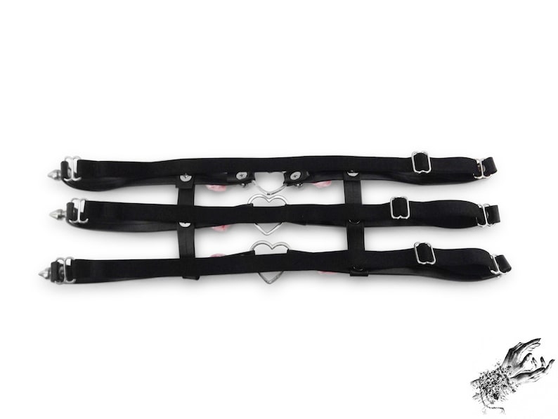 Rear view of a black thigh garter showing 3 black satin elastic straps with adjustable sliders for a comfortable and versatile fit. Design features silver heart rings, pink satin roses and silver spike studs for an edgy yet kawaii look. Perfect for alternative fashion styles including pastel goth, Harajuku fashion and coquette aesthetics, as well as cosplay and BDSM communities. Displayed on a white background.