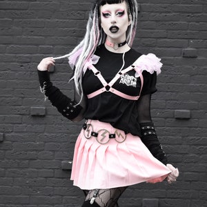 Modelled photo of the pink ruffled harness bra, styled with a matching black and pink heart ring choker, black and white graphic t-shirt and pink pleated skirt for a pastel goth fashion aesthetic. The adjustable shoulder and bust straps at the back ensures a comfortable, versatile fit for all body types and sizes. Perfect for alternative fashion styles including pastel goth, kawaii and e-girl style, as well as BDSM inspired outfits, burlesque costumes, festival fashion and ravewear.