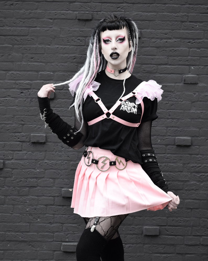 Modelled photo of the pink ruffled harness bra, styled with a matching black and pink heart ring choker, black and white graphic t-shirt and pink pleated skirt for a pastel goth fashion aesthetic. The adjustable shoulder and bust straps at the back ensures a comfortable, versatile fit for all body types and sizes. Perfect for alternative fashion styles including pastel goth, kawaii and e-girl style, as well as BDSM inspired outfits, burlesque costumes, festival fashion and ravewear.
