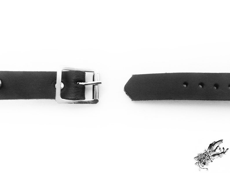 A photo of the back of the black leather O ring wristband - one end has a silver coloured buckle, and the other end has multiple holes to fit 16cm to 21cm wrist circumference.