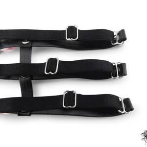 Rear view of a black thigh garter showing 3 black satin elastic straps with adjustable sliders for a comfortable and versatile fit. Design features silver heart rings, pink satin roses and silver spike studs for an edgy yet kawaii look. Perfect for alternative fashion styles including pastel goth, Harajuku fashion and coquette aesthetics, as well as cosplay and BDSM communities. Displayed on a white background.