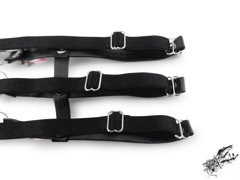 Rear view of a black thigh garter showing 3 black satin elastic straps with adjustable sliders for a comfortable and versatile fit. Design features silver heart rings, pink satin roses and silver spike studs for an edgy yet kawaii look. Perfect for alternative fashion styles including pastel goth, Harajuku fashion and coquette aesthetics, as well as cosplay and BDSM communities. Displayed on a white background.