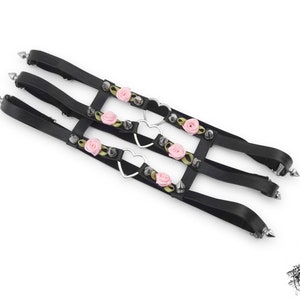Black thigh garter featuring 3 horizontal straps with central silver heart ring hardware, pink satin roses and silver spike studs either side. Adjustable elastic back ensures a comfortable and versatile fit. Perfect for alternative fashion styles including pastel goth, Harajuku fashion and coquette aesthetics, as well as cosplay and BDSM communities. Displayed on a white background.