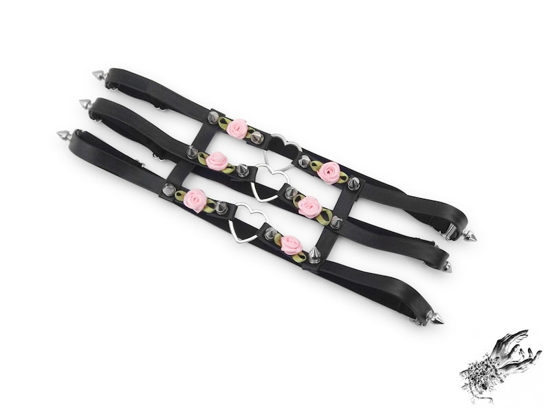 Black thigh garter featuring 3 horizontal straps with central silver heart ring hardware, pink satin roses and silver spike studs either side. Adjustable elastic back ensures a comfortable and versatile fit. Perfect for alternative fashion styles including pastel goth, Harajuku fashion and coquette aesthetics, as well as cosplay and BDSM communities. Displayed on a white background.