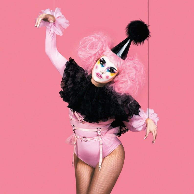 Modelled photo of the pink O ring garter belt, styled with curly pink hair buns, pastel clown make-up, pink bodysuit and black ruffled capelet for a kawaii clown aesthetic. The adjustable elastic waist and suspender straps and faux leather hip straps provide a versatile, comfortable fit for all body types and sizes. Perfect for alternative fashion styles including pastel goth, Harajuku fashion and e-girl style, as well as BDSM outfits, burlesque costumes, festival fashion and ravewear.