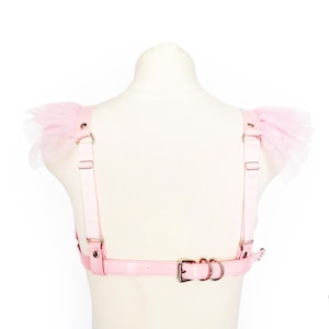 Rear view of a pink vegan leather ruffled harness, featuring pastel pink faux leather straps, durable silver hardware, ruffled tulle shoulder accents and an adjustable buckle strap for a secure fit. Elasticated shoulder straps with sliding adjusters provide added comfort and flexibility, making it a versatile accessory for pastel goth, Japanese street fashion, burlesque costumes, festival or BDSM-inspired outfits. Displayed on a white mannequin bust to show the flattering, body conforming fit.