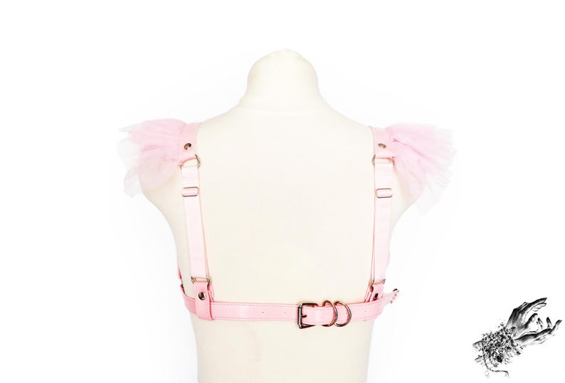 Rear view of a pink vegan leather ruffled harness, featuring pastel pink faux leather straps, durable silver hardware, ruffled tulle shoulder accents and an adjustable buckle strap for a secure fit. Elasticated shoulder straps with sliding adjusters provide added comfort and flexibility, making it a versatile accessory for pastel goth, Japanese street fashion, burlesque costumes, festival or BDSM-inspired outfits. Displayed on a white mannequin bust to show the flattering, body conforming fit.