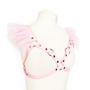 Adjustable pink vegan leather harness, featuring silver rivets and O rings, and pink ruffled tulle shoulder accents. This pastel pink vegan leather harness combines durability and bold design, perfect for Japanese street fashion, pastel goth and alternative fashion enthusiasts. Also suitable for BDSM communities seeking a stylish, playproof chest harness. Great for cosplay, Halloween, or festival outfits, the harness offers versatility and a comfortable, customizable fit for various body types.