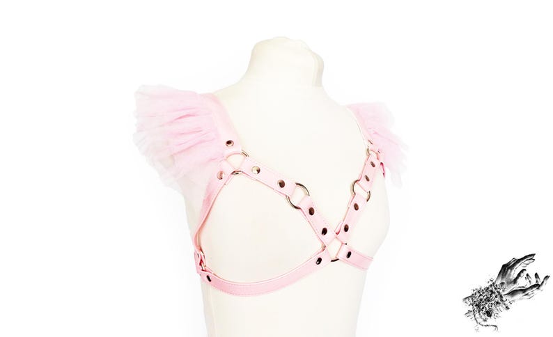 Adjustable pink vegan leather harness, featuring silver rivets and O rings, and pink ruffled tulle shoulder accents. This pastel pink vegan leather harness combines durability and bold design, perfect for Japanese street fashion, pastel goth and alternative fashion enthusiasts. Also suitable for BDSM communities seeking a stylish, playproof chest harness. Great for cosplay, Halloween, or festival outfits, the harness offers versatility and a comfortable, customizable fit for various body types.