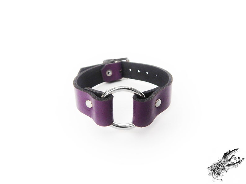 Purple leather wristband with a silver O ring in the center, and two silver coloured flat shaped rivets holding the purple leather and O ring in place. There is a silver buckle at the back with multiple holes to fit 16cm to 21cm wrist circumference.