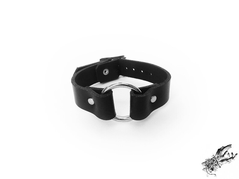 Black leather wristband with a silver O ring in the center, and two silver coloured flat shaped rivets holding the black leather and O ring in place. There is a silver buckle at the back with multiple holes to fit 16cm to 21cm wrist circumference.