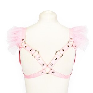 Adjustable pink vegan leather harness, featuring silver rivets and O rings, and pink ruffled tulle shoulder accents. This pastel pink vegan leather harness combines durability and bold design, perfect for Japanese street fashion, pastel goth and alternative fashion enthusiasts. Also suitable for BDSM communities seeking a stylish, playproof chest harness. Great for cosplay, Halloween, or festival outfits, the harness offers versatility and a comfortable, customizable fit for various body types.