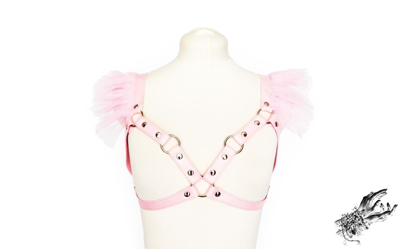 Adjustable pink vegan leather harness, featuring silver rivets and O rings, and pink ruffled tulle shoulder accents. This pastel pink vegan leather harness combines durability and bold design, perfect for Japanese street fashion, pastel goth and alternative fashion enthusiasts. Also suitable for BDSM communities seeking a stylish, playproof chest harness. Great for cosplay, Halloween, or festival outfits, the harness offers versatility and a comfortable, customizable fit for various body types.