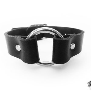 Close-up photo of a black leather wristband with a silver coloured O ring in the center, and two silver coloured flat shaped rivets holding the black leather and silver O ring in place.