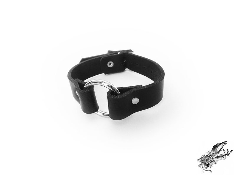 Black leather wristband with a silver coloured O ring in the center, and two silver coloured flat shaped rivets holding the black leather and silver O ring in place.