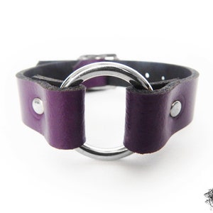 Close-up photo of a purple leather wristband with a silver coloured O ring in the center, and two silver coloured flat shaped rivets holding the purple leather and silver O ring in place.