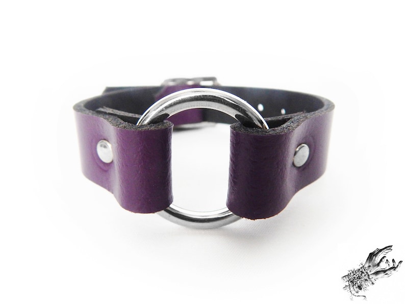 Close-up photo of a purple leather wristband with a silver coloured O ring in the center, and two silver coloured flat shaped rivets holding the purple leather and silver O ring in place.
