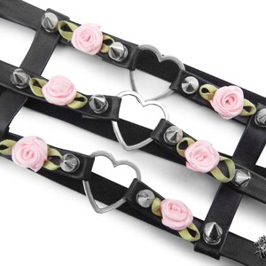 Close-up of a black leather thigh garter featuring 3 horizontal straps with central silver heart ring hardware, pink satin roses and silver spike studs either side. Adjustable elastic back ensures a comfortable and versatile fit. Perfect for alternative fashion styles including pastel goth, Harajuku fashion and coquette aesthetics, as well as cosplay and BDSM communities. Displayed on a white background.