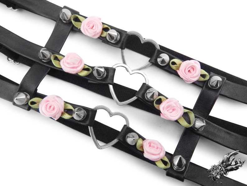 Close-up of a black leather thigh garter featuring 3 horizontal straps with central silver heart ring hardware, pink satin roses and silver spike studs either side. Adjustable elastic back ensures a comfortable and versatile fit. Perfect for alternative fashion styles including pastel goth, Harajuku fashion and coquette aesthetics, as well as cosplay and BDSM communities. Displayed on a white background.