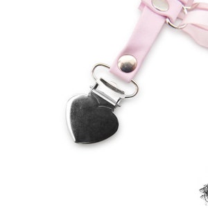 Close-up of a pink corset lacing thigh garter, showing the silver metal rivets securely attaching the silver heart shaped suspender clips, perfect for fastening stockings and knee high socks. The metal heart suspender clips ensure a comfortable and versatile fit, blending style and functionality effortlessly. A perfect alternative thigh garter for brides and weddings, as well as fashion styles such as pastel goth, kawaii and Japanese fashion. Photographed on a white background.