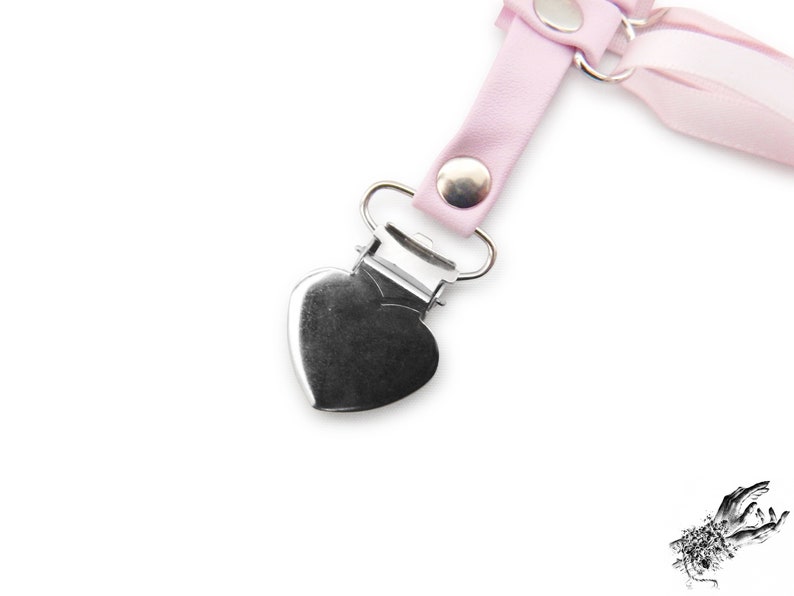 Close-up of a pink corset lacing thigh garter, showing the silver metal rivets securely attaching the silver heart shaped suspender clips, perfect for fastening stockings and knee high socks. The metal heart suspender clips ensure a comfortable and versatile fit, blending style and functionality effortlessly. A perfect alternative thigh garter for brides and weddings, as well as fashion styles such as pastel goth, kawaii and Japanese fashion. Photographed on a white background.