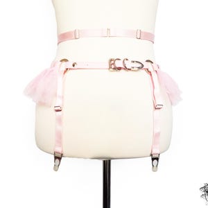 Rear view of a pink vegan leather garter belt, featuring ruffled tulle accents at the hips, durable silver hardware, and an adjustable buckle strap at the hips for a secure fit. The elasticated waist strap with sliding adjuster and hook and eye fastening provides added comfort and flexibility, making it a versatile accessory for pastel goth and kawaii fashion, burlesque costumes, festival or BDSM-inspired outfits. Displayed on a white mannequin bust to show the flattering, body conforming fit.