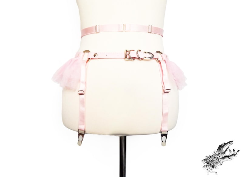 Rear view of a pink vegan leather garter belt, featuring ruffled tulle accents at the hips, durable silver hardware, and an adjustable buckle strap at the hips for a secure fit. The elasticated waist strap with sliding adjuster and hook and eye fastening provides added comfort and flexibility, making it a versatile accessory for pastel goth and kawaii fashion, burlesque costumes, festival or BDSM-inspired outfits. Displayed on a white mannequin bust to show the flattering, body conforming fit.