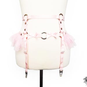 Adjustable pink vegan leather garter belt crafted for both functionality and style, catering to alternative fashion enthusiasts and BDSM communities alike. Featuring a central silver O ring, pink satin bow details, ruffled tulle accents at the side of the hips and pink elastic suspender straps, it’s a versatile garter belt that complements pastel goth and Japanese fashion, burlesque costumes or festival outfits. Displayed on a white mannequin bust to show the flattering, body conforming fit.