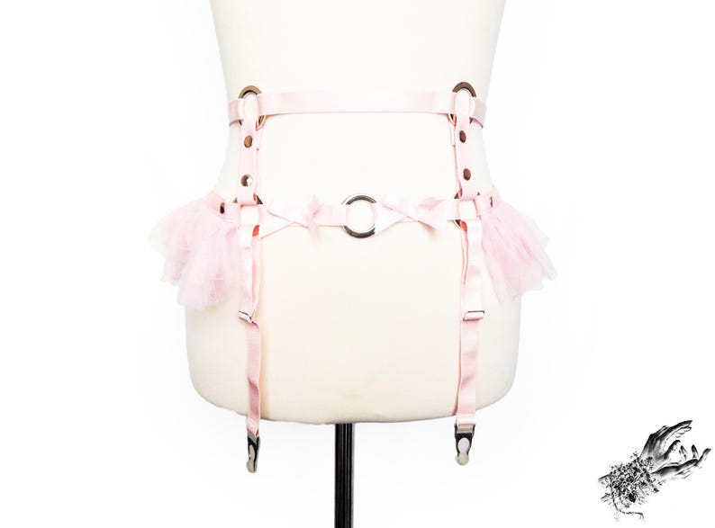 Adjustable pink vegan leather garter belt crafted for both functionality and style, catering to alternative fashion enthusiasts and BDSM communities alike. Featuring a central silver O ring, pink satin bow details, ruffled tulle accents at the side of the hips and pink elastic suspender straps, it’s a versatile garter belt that complements pastel goth and Japanese fashion, burlesque costumes or festival outfits. Displayed on a white mannequin bust to show the flattering, body conforming fit.