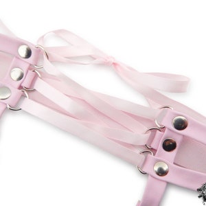 Close-up of a pink corset lacing thigh garter, showing the silver metal rivets securely attaching the pink satin ribbon ties in a criss-cross corset lacing style. The adjustable ribbon ties ensure a comfortable and versatile fit, blending style and functionality effortlessly. A perfect alternative thigh garter for brides and weddings, as well as alternative fashion styles such as pastel goth, kawaii and Japanese fashion. Displayed on a white background.