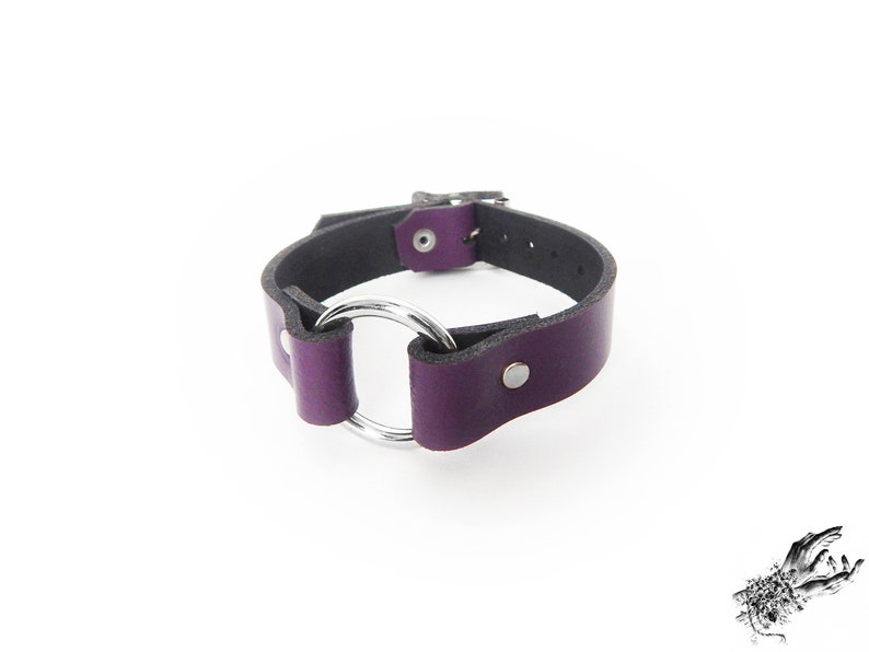 Purple leather wristband with a silver coloured O ring in the center, and two silver coloured flat shaped rivets holding the purple leather and silver O ring in place.