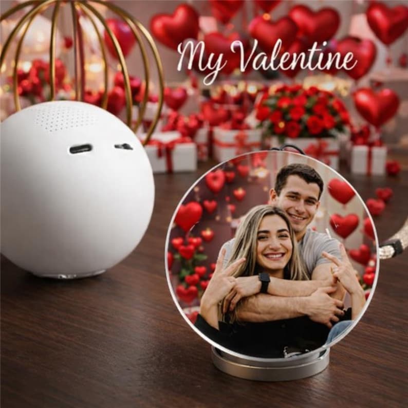 May include: A white, spherical speaker with a small, round design, next to a circular photo frame with a couple embracing. The photo frame is on a silver base. The background features red heart-shaped balloons and the text "My Valentine".