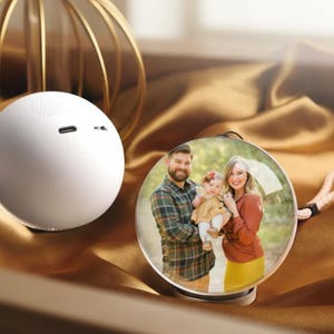 May include: A white, spherical speaker with a small, round design, next to a clear, round photo frame. The frame displays a family portrait. The background is a draped, golden-brown fabric.