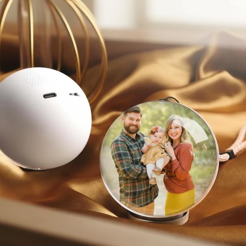 May include: A white, spherical speaker with a small, round design, next to a clear, round photo frame. The frame displays a family portrait. The background is a draped, golden-brown fabric.