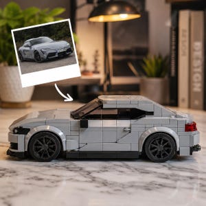 Op de afbeelding: Een grijs bouwpakketmodel van een sportwagen in zijaanzicht, met zwarte wielen, een lage carrosserie en een transparante voorruit. Boven het model staat een vierkante foto van een grijze sportwagen op een ondergrond van grind.