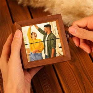 May include: A square wooden photo frame with a rotating mechanism. The frame contains a photograph of a couple dancing. The frame is held by a person's hands, set against a wooden surface.