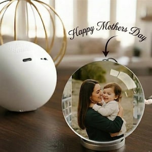 May include: A clear, spherical photo ornament featuring a mother and child. The ornament sits on a wooden surface, with a white speaker and a gold decorative sphere in the background. The text "Happy Mothers Day" is above the ornament.