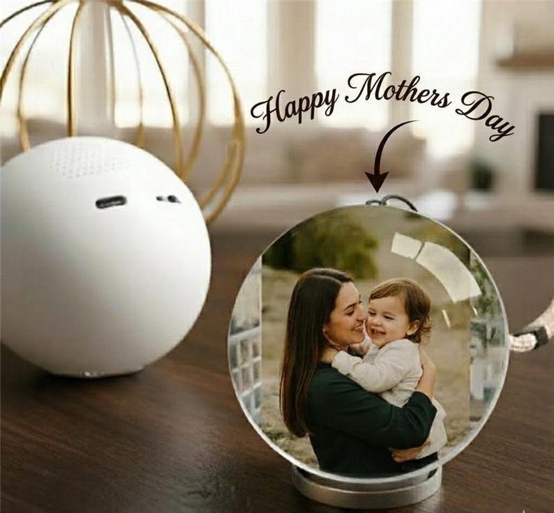May include: A clear, spherical photo ornament featuring a mother and child. The ornament sits on a wooden surface, with a white speaker and a gold decorative sphere in the background. The text "Happy Mothers Day" is above the ornament.