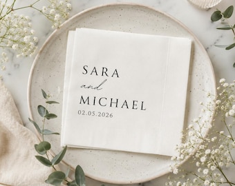 May include: White paper napkins with the names "SARA and MICHAEL" and the date "02.05.2026" printed in black. The napkins are on a white plate, surrounded by greenery and a lit candle.