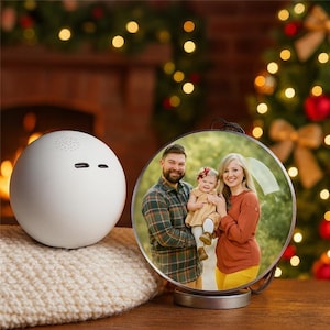 Op de afbeelding: Een witte, bolvormige luidspreker met twee horizontale sleuven en een luidsprekerrooster ligt op een crèmekleurig gebreid oppervlak. Een ronde, heldere fotolijst toont een familieportret. De achtergrond toont een open haard en een versierde kerstboom.