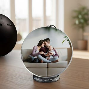 May include: A black, spherical speaker sits on a wooden surface next to a clear, round photo display. The display shows a family portrait and has the text "Moments Made Timeless" on the base.