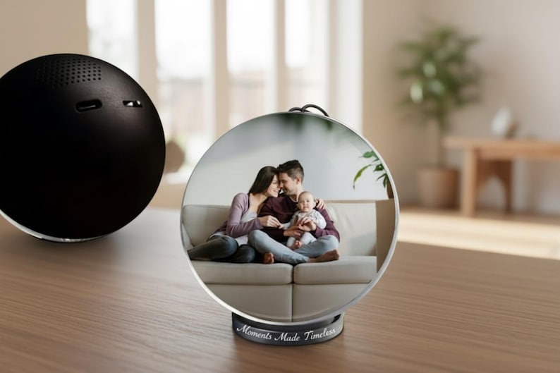 May include: A black, spherical speaker sits on a wooden surface next to a clear, round photo display. The display shows a family portrait and has the text "Moments Made Timeless" on the base.