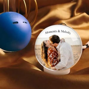 May include: A blue spherical speaker beside a clear glass ornament. The ornament features a photo of a kissing couple, with the text "Moments & Melody". The background is a brown, silky fabric.