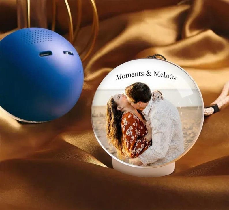 May include: A blue spherical speaker beside a clear glass ornament. The ornament features a photo of a kissing couple, with the text "Moments & Melody". The background is a brown, silky fabric.