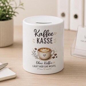 May include: A white ceramic money box featuring a horizontal coin slot at the top. The front displays the text "Kaffee KASSE" above an illustration of a coffee cup with latte art, followed by the text "Ohne Kaffee LÄUFT HIER GAR NICHTS."