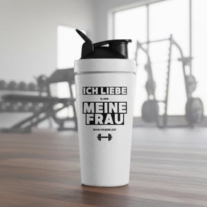 Könnte beinhalten: Weißer Shaker mit schwarzem Deckel und Klappverschluss. Der Shaker trägt den schwarzen Text "ICH LIEBE ES, WENN MEINE FRAU MICH INS GYM GEHEN LÄSST" und ein Hantel-Symbol. Ideal für Fitnessstudio-Besuche und Sport.