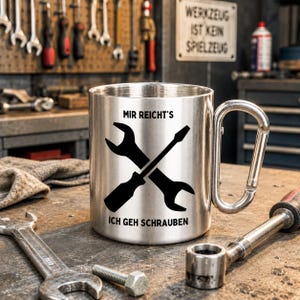 Pode incluir: Uma caneca de aço inoxidável prateada com uma pega em forma de mosquetão. A caneca apresenta um gráfico preto de uma chave inglesa e uma chave de fendas cruzadas, com o texto alemão "MIR REICHT'S" acima e "ICH GEH SCHRAUBEN" abaixo.
